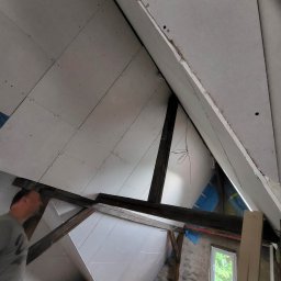 MatBud - Montaż płyt karton gips na poddaszu, widoczne drewniane belki konstrukcyjne, fragment okna i wiadro z odpadami po cięciu płyt.