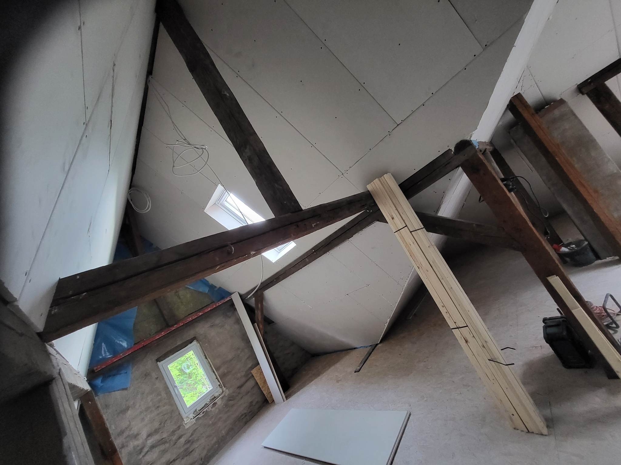 Montaż płyt kartonowo-gipsowych na poddaszu, widoczne drewniane belki konstrukcyjne, okno dachowe i przygotowane płyty na podłodze.