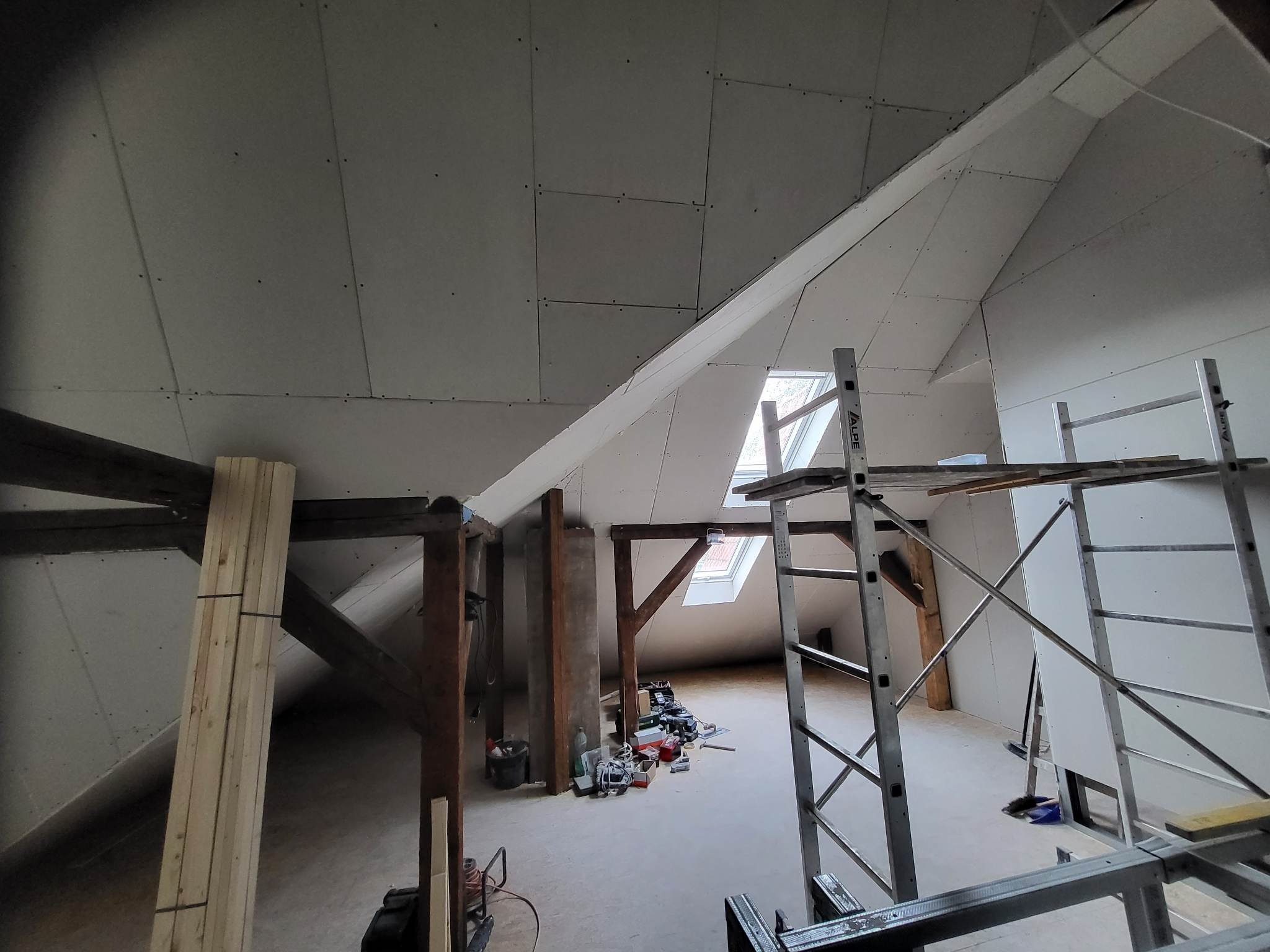 Poddasze w trakcie prac wykończeniowych: widoczne płyty kartonowo-gipsowe na skośnym suficie, drewniane belki konstrukcyjne, aluminiowe rusztowanie oraz narzędzia rozłożone na podłodze.