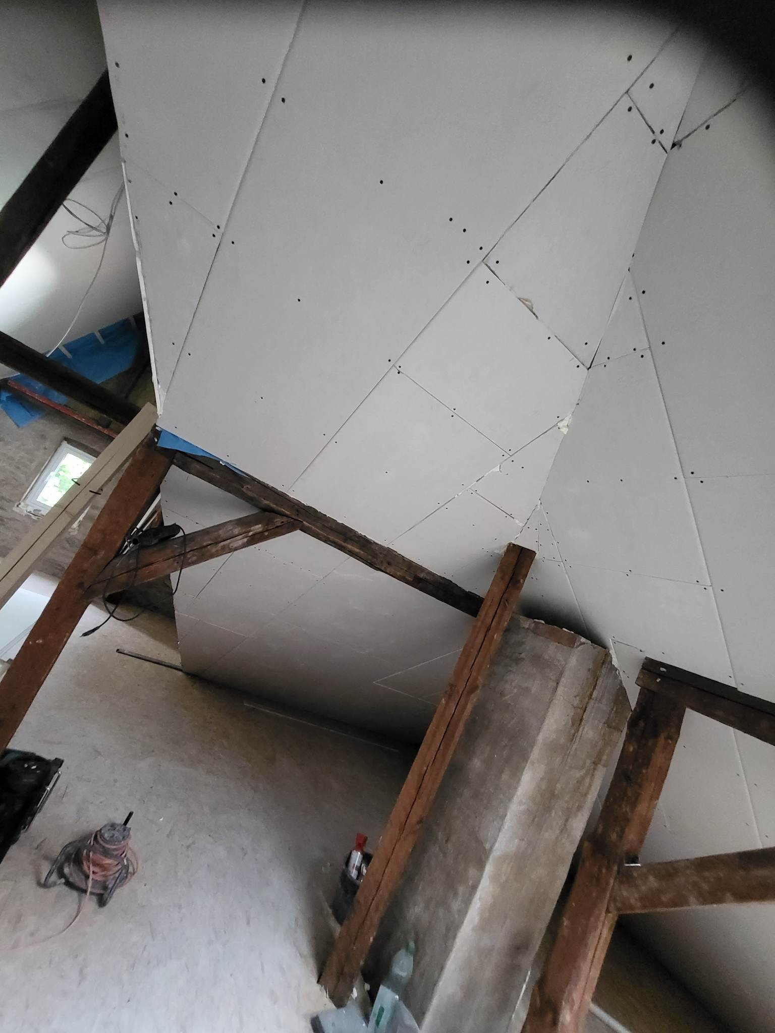 Skośny sufit na poddaszu w trakcie montażu płyt kartonowo-gipsowych, widoczne drewniane belki konstrukcyjne i narzędzia na podłodze.