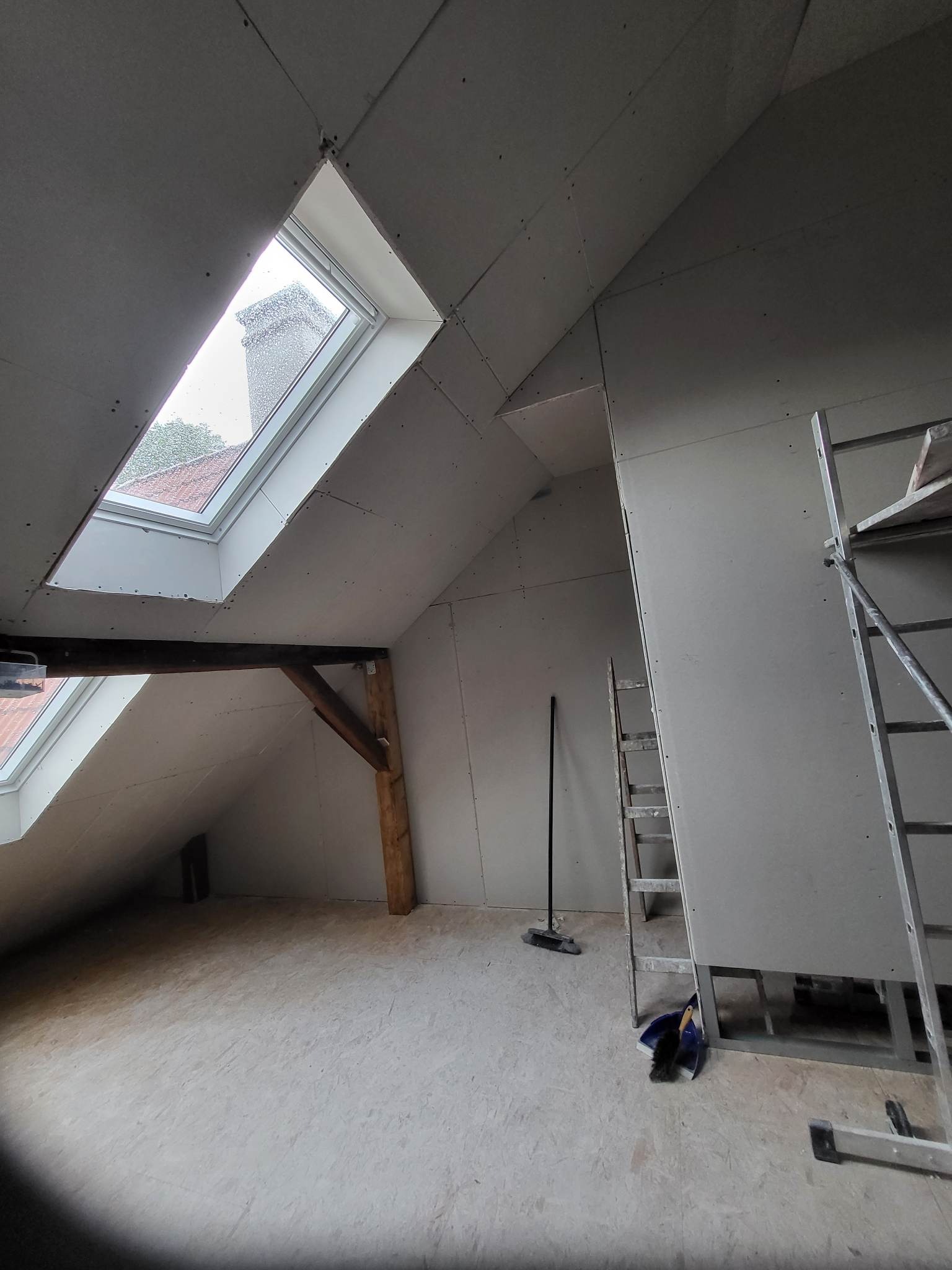 Poddasze w trakcie wykańczania płytami kartonowo-gipsowymi, widoczne okna dachowe, drewniane belki konstrukcyjne, drabina i narzędzia do sprzątania.