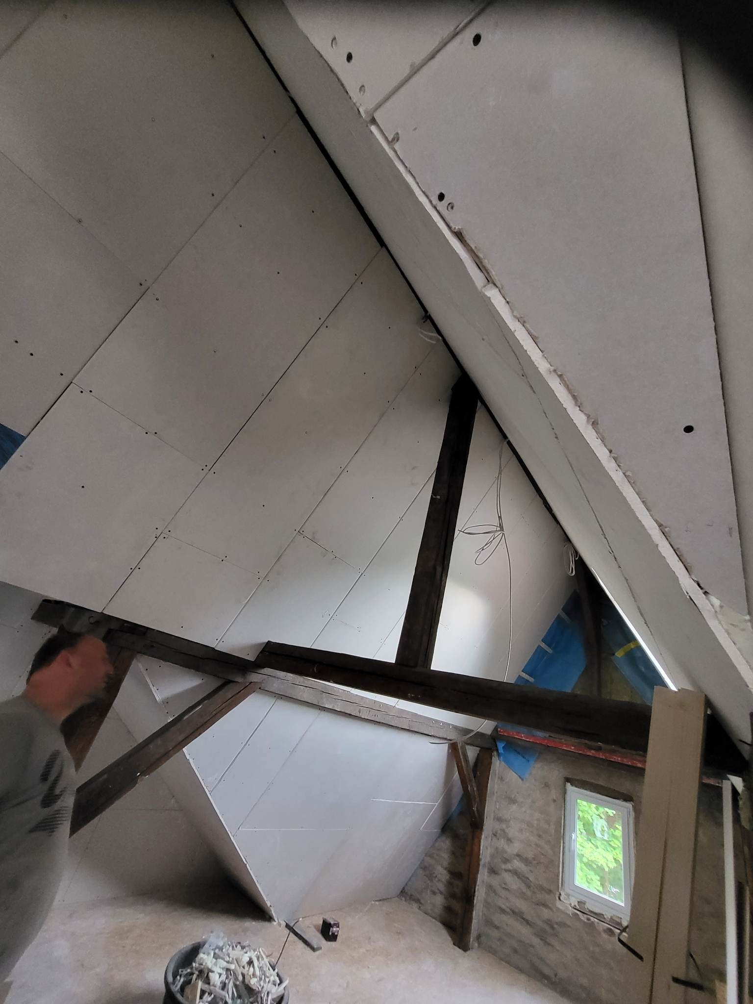 Montaż płyt karton gips na poddaszu, widoczne drewniane belki konstrukcyjne, fragment okna i wiadro z odpadami po cięciu płyt.