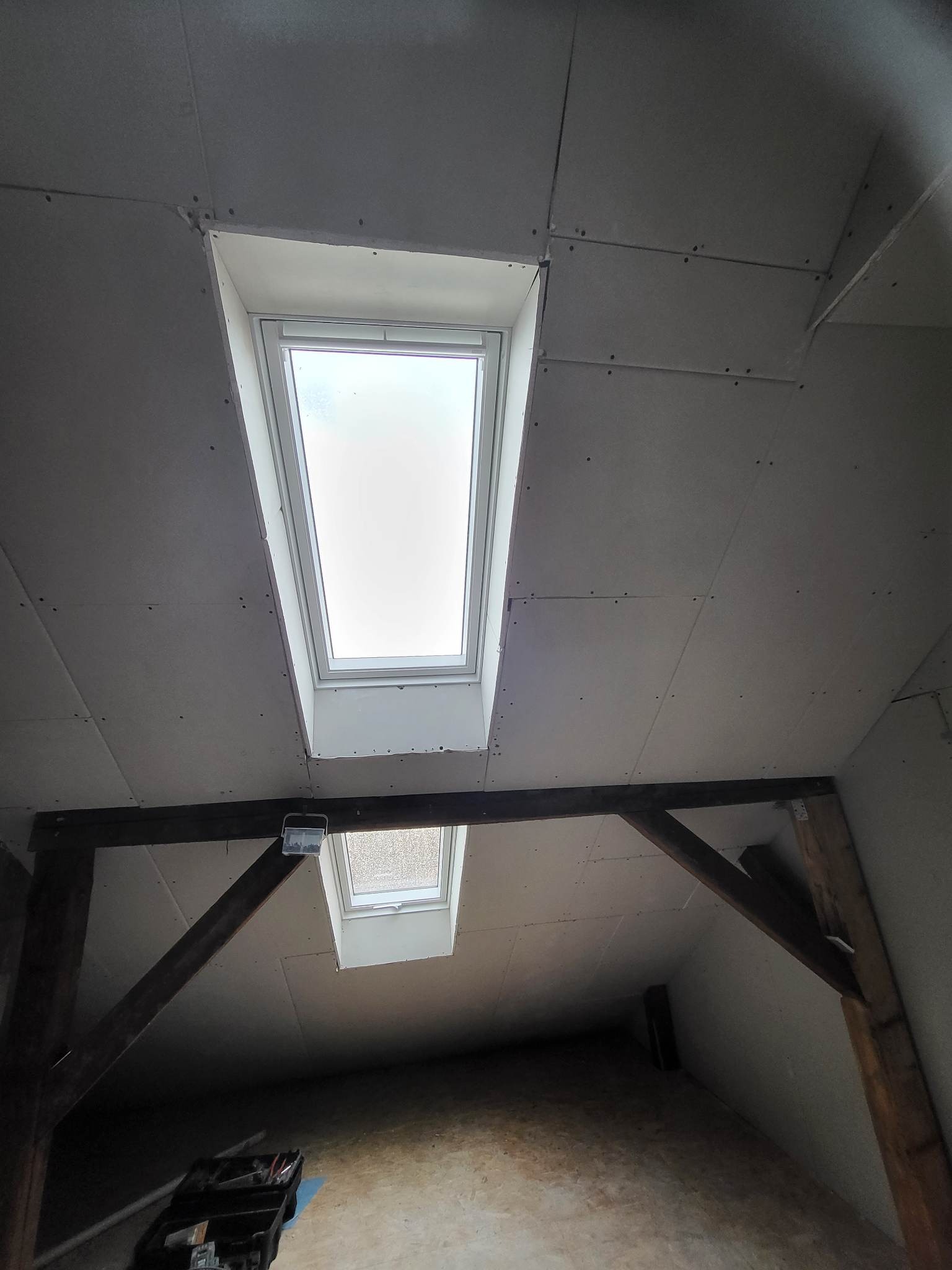 Montaż płyt kartonowo-gipsowych na poddaszu z oknami dachowymi, widoczne drewniane belki konstrukcyjne i skrzynka z narzędziami na podłodze.