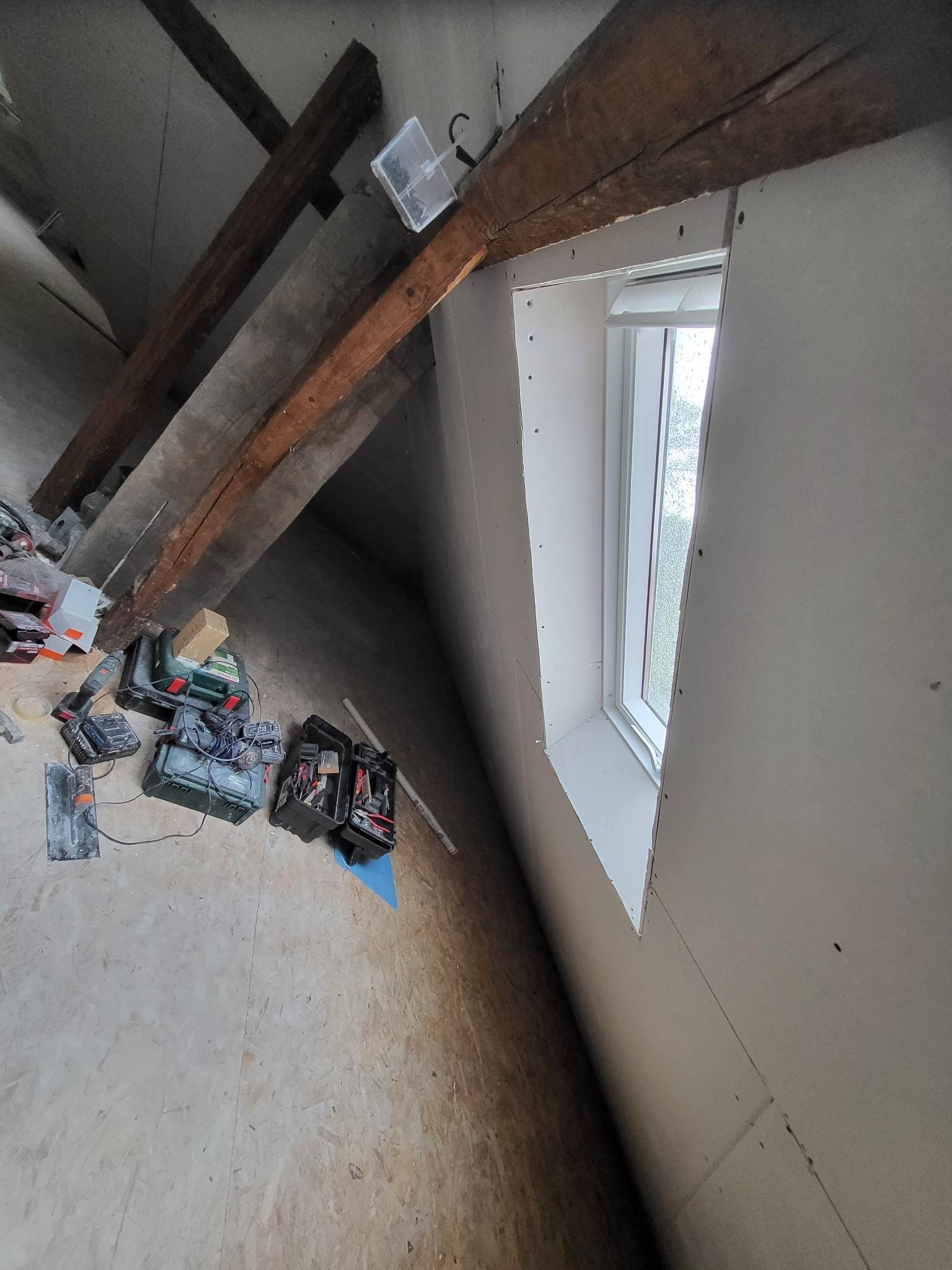 Montaż płyty karton gips wokół okna na poddaszu, widoczne drewniane belki stropowe, narzędzia i skrzynki narzędziowe na podłodze z płyt OSB.