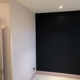 Kamil Noga - Pokój po odświeżeniu: ściana w kolorze pudrowego różu sąsiaduje z czarną ścianą, listwy przypodłogowe w odcieniu różu, na podłodze leży kabel i puszka farby.