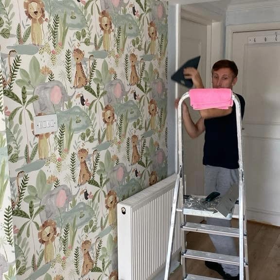 Mężczyzna na drabinie wygładza raklą tapetę w dziecięcym stylu z motywami zwierząt, widoczny grzejnik i włącznik światła na ścianie.
