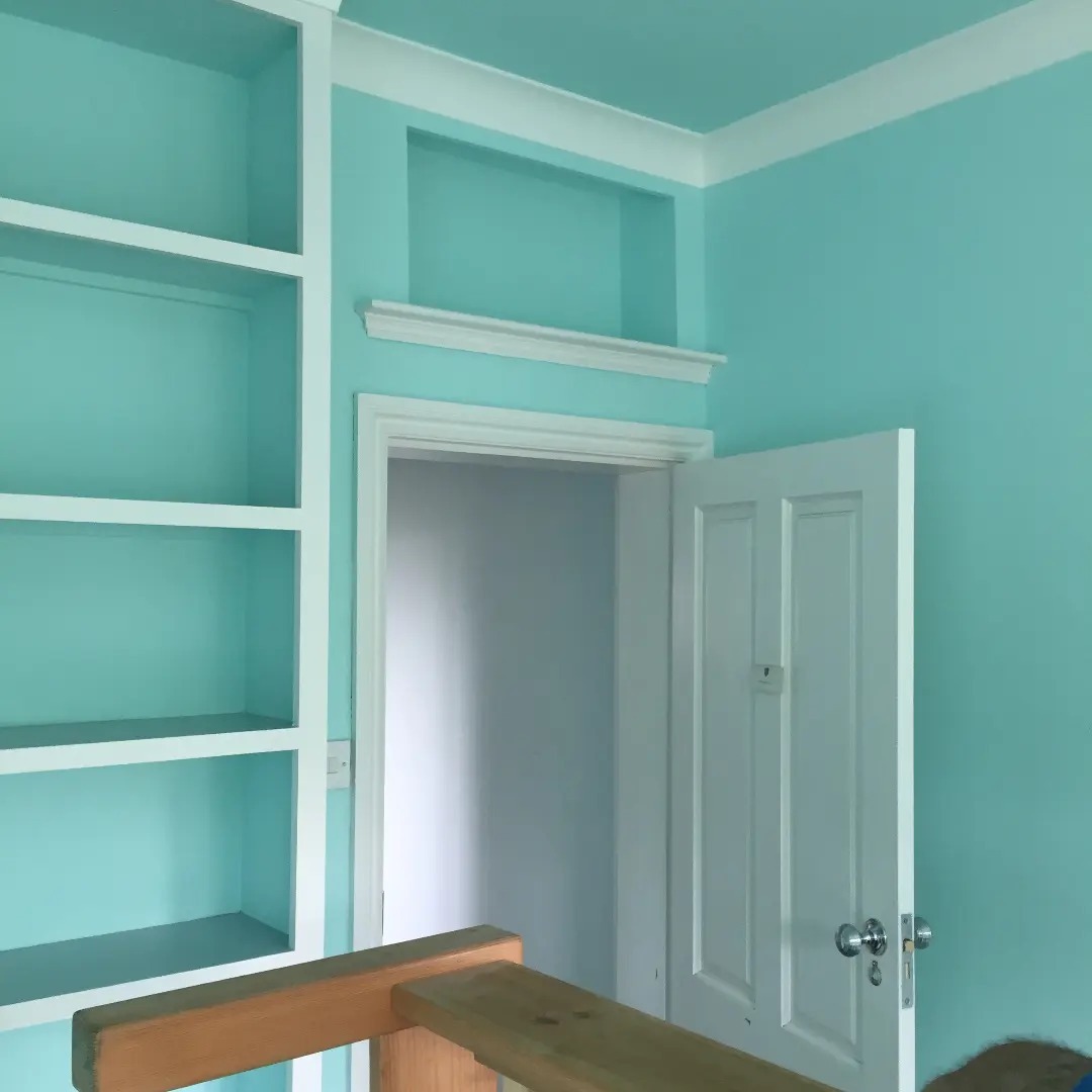 Pomieszczenie pomalowane na błękitno z białymi listwami, otwartymi półkami i białymi drzwiami z srebrną klamką, widoczny fragment rusztowania.