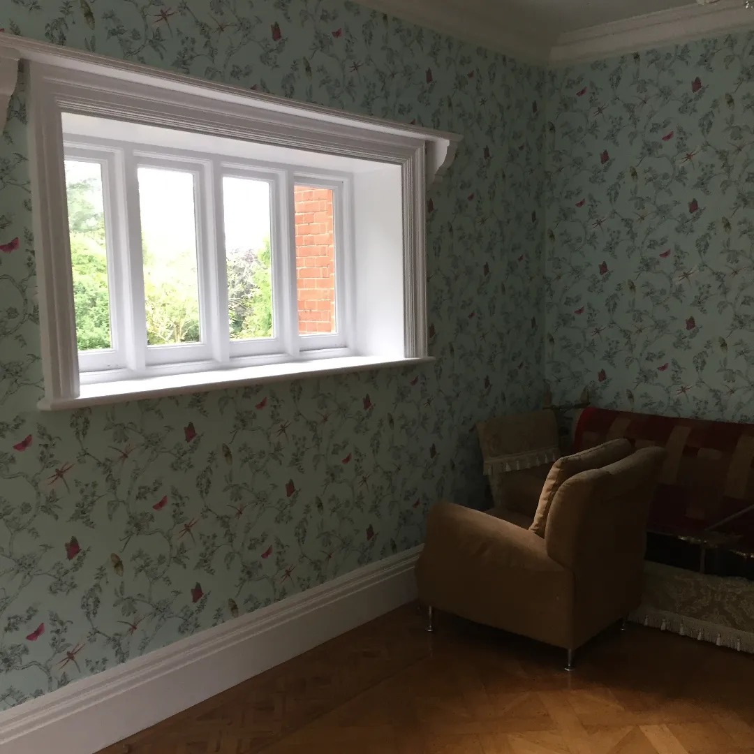 Pokój z jasną tapetą w motywy roślinne i owadzie, białe okno z widokiem na zieleń, parkiet na podłodze, fotel i sofa w rogu pokoju.