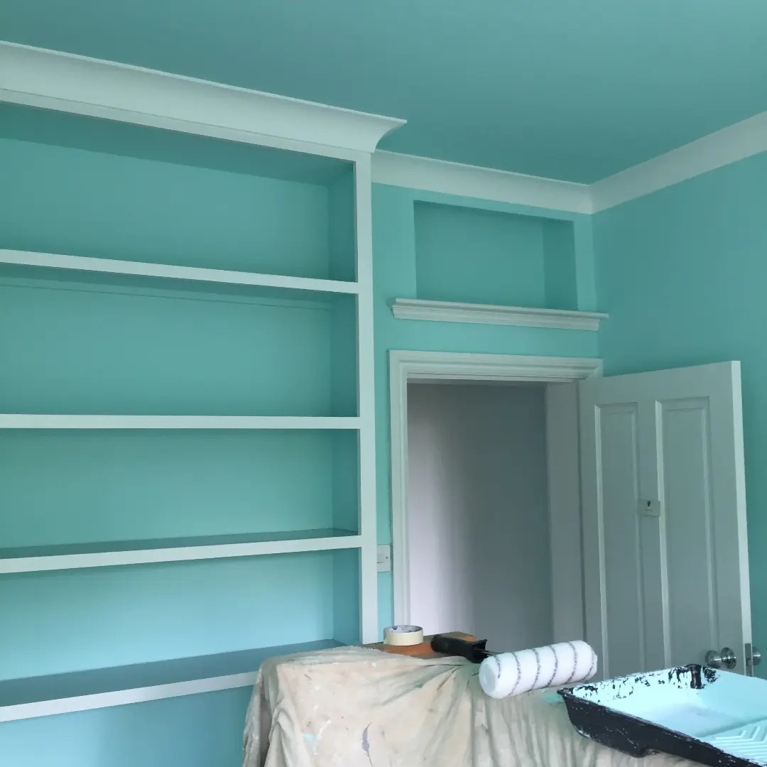 Świeżo pomalowane na turkusowo wnętrze pokoju z białymi półkami, listwami i drzwiami; widoczny wałek malarski i kuweta z farbą na zabezpieczonym stole.