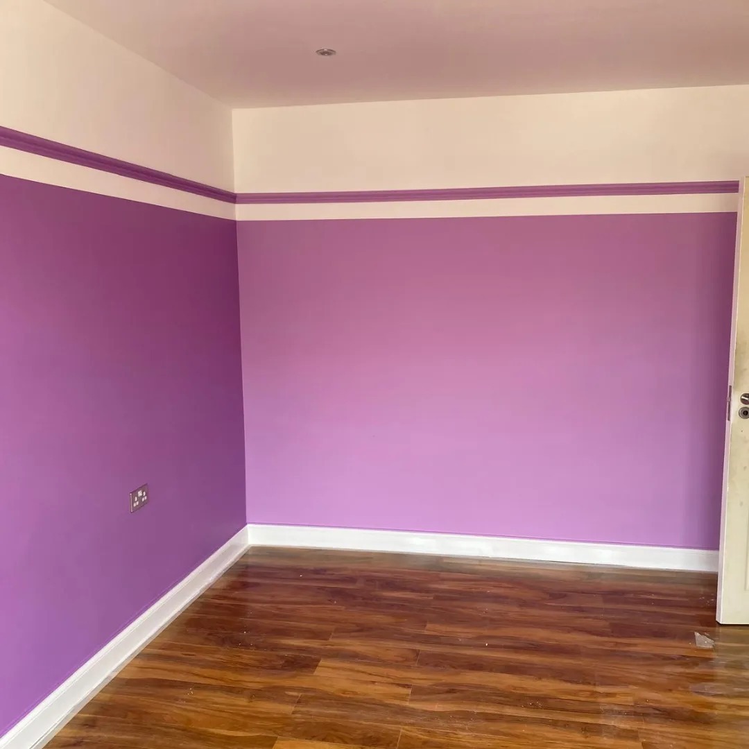 Pokój po remoncie z fioletowymi ścianami, białym pasem dekoracyjnym pod sufitem i brązową podłogą imitującą drewno. Widoczny fragment drzwi i gniazdko elektryczne.