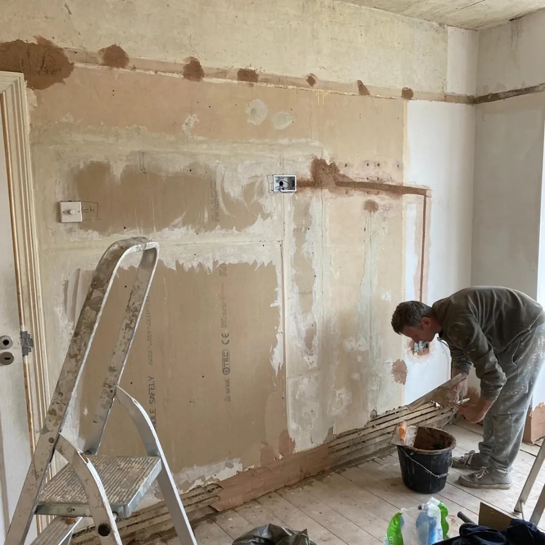 Mężczyzna szpachluje ścianę w trakcie remontu, widoczna drabina, puszki elektryczne i przygotowana ściana z płyt karton gips.