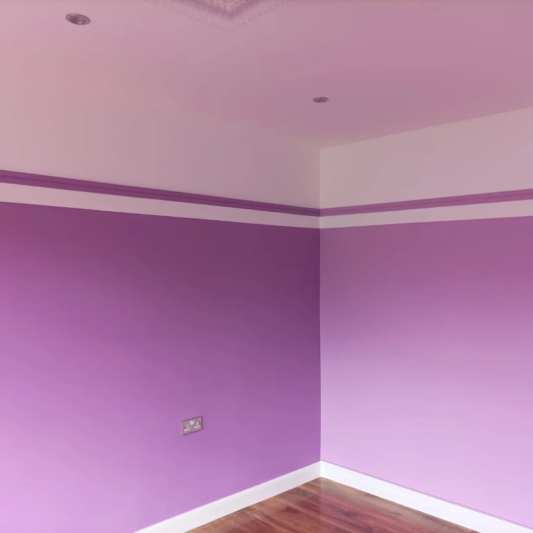 Pomalowane na fioletowo ściany w rogu pokoju z drewnianą podłogą, widoczne gniazdko elektryczne i biały pas pod sufitem.