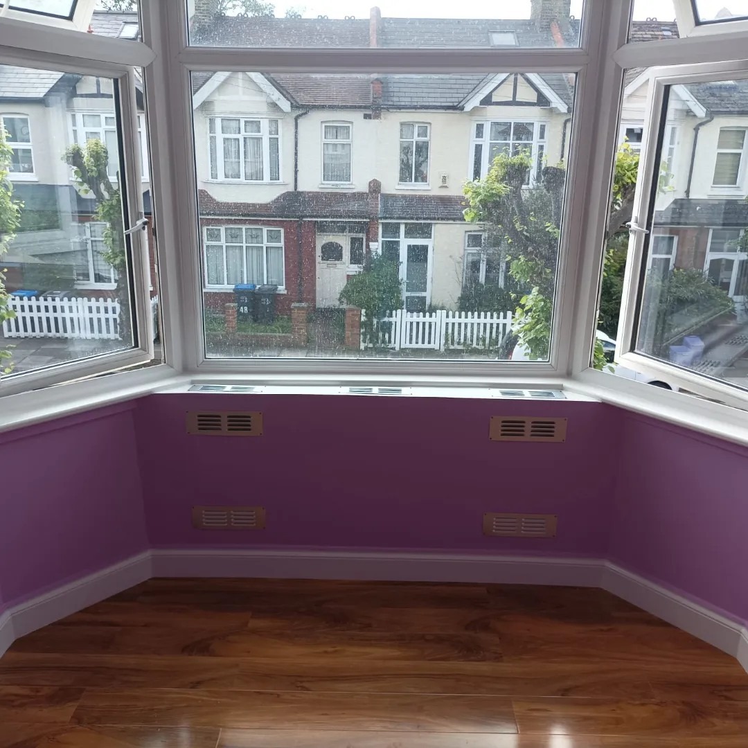 Wnętrze pokoju z wykończoną wnęką okienną w odcieniach fioletu, z drewnianą podłogą i widokiem na sąsiednie domy przez otwarte okna.