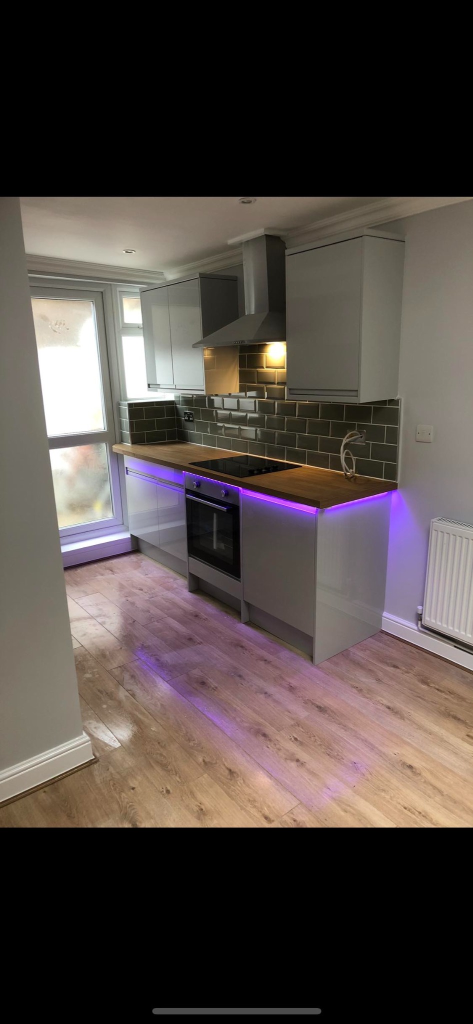 Wykończona kuchnia w szarościach z drewnianym blatem, podświetleniem LED w kolorze fioletowym pod szafkami i ciemnozielonymi płytkami na ścianie.