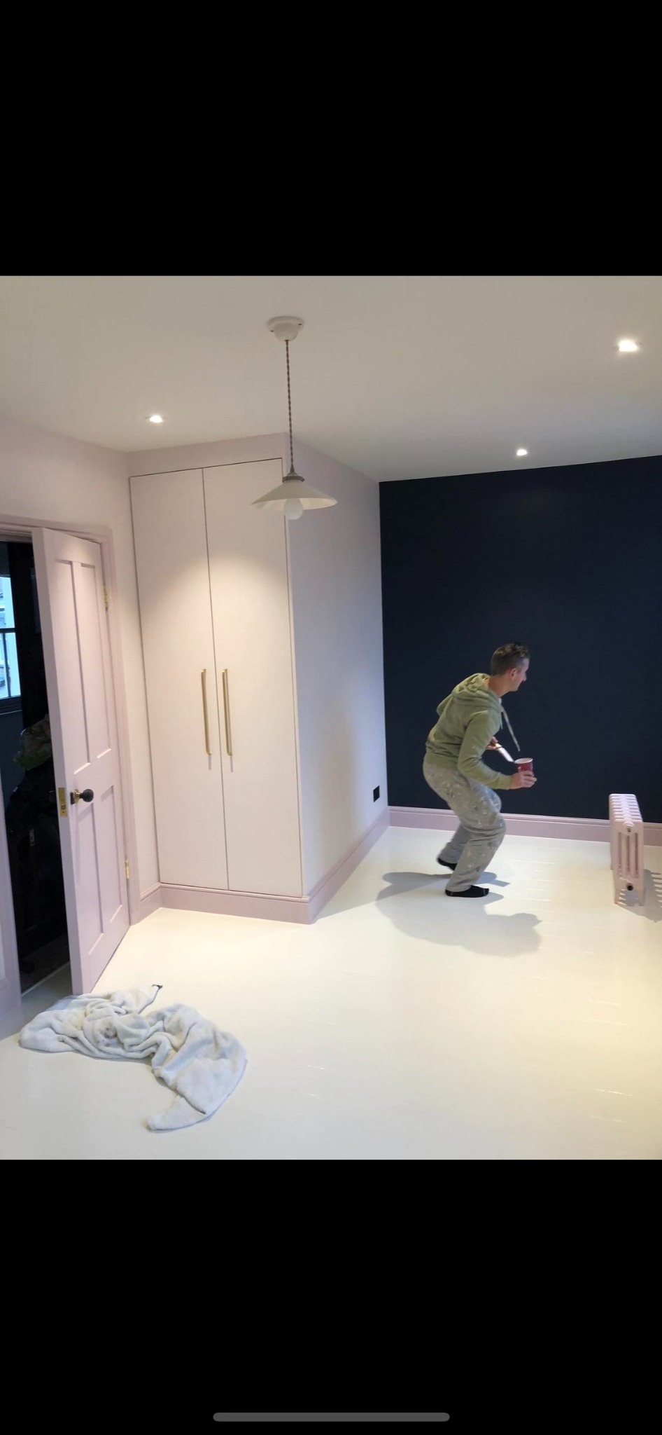 Mężczyzna w trakcie malowania ściany na granatowo w pokoju z białymi podłogami, różowymi detalami i grzejnikiem; widoczne ślady farby na podłodze i ubraniu malarza.