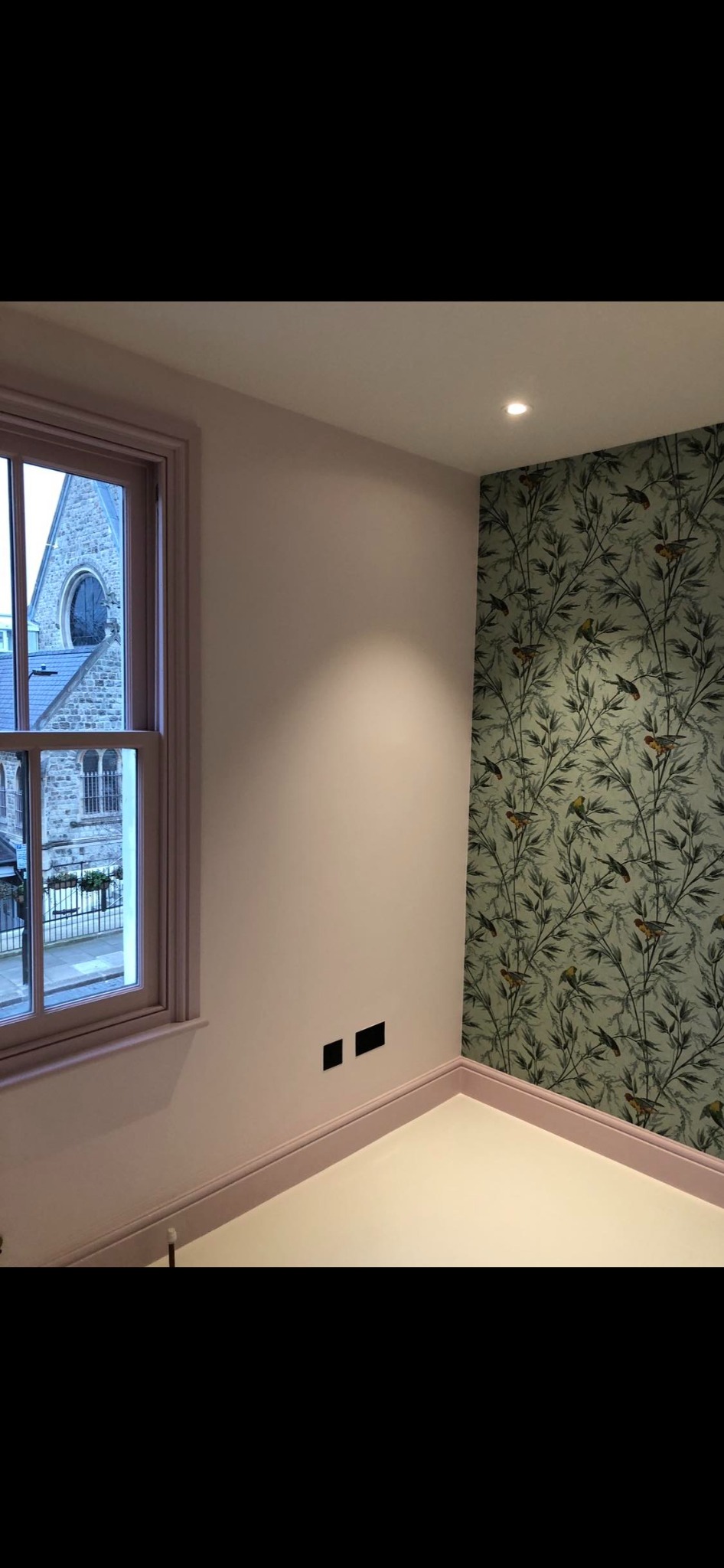 Pokój po remoncie: ściana pomalowana na jasny róż, fragment ściany z tapetą w motywy roślinne i ptasie, okno z widokiem na kamienny budynek.