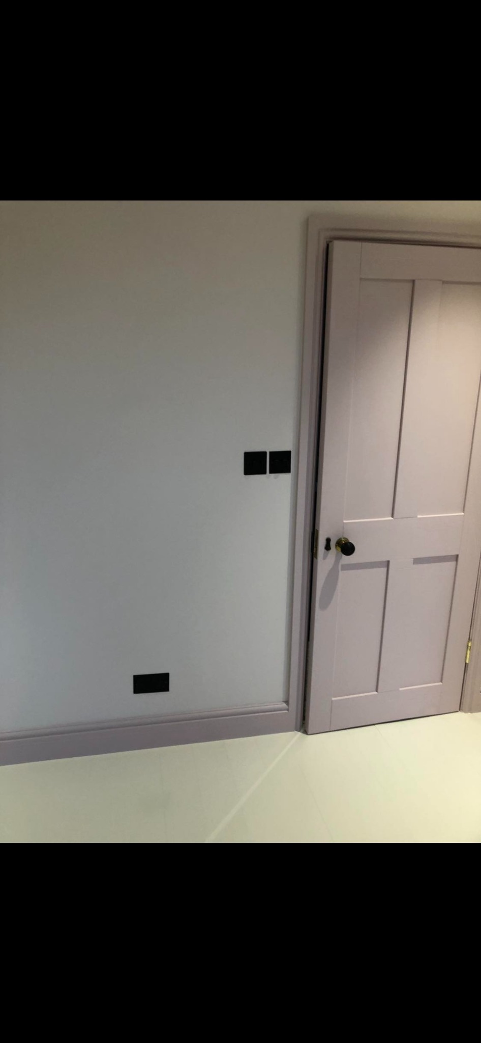 Ściana pomalowana na jasny odcień z czarnymi włącznikami światła i listwą przypodłogową w kolorze liliowym, obok liliowe drzwi z czarną klamką.