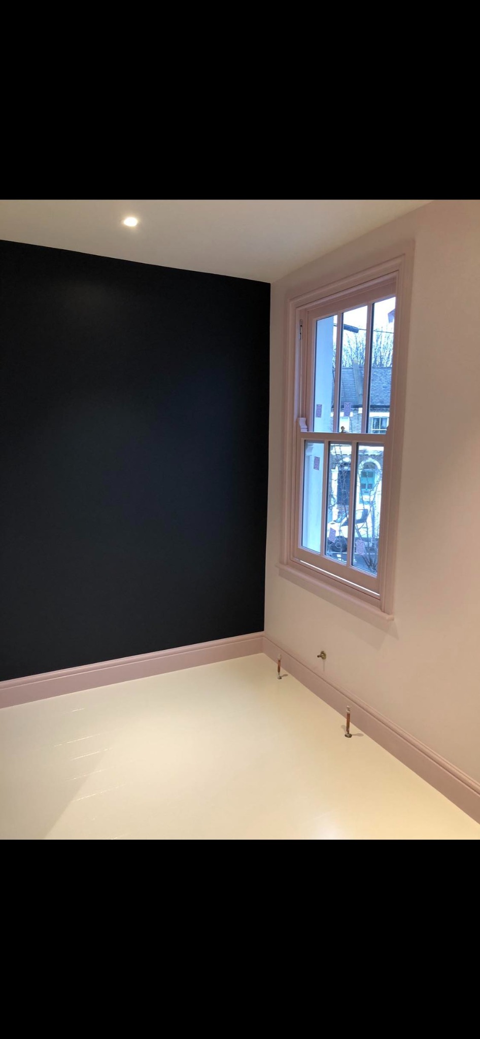 Pomieszczenie po malowaniu: czarna ściana kontrastuje z jasnoróżowymi listwami przypodłogowymi i ramą okienną, widoczne wystające rurki instalacji grzewczej.