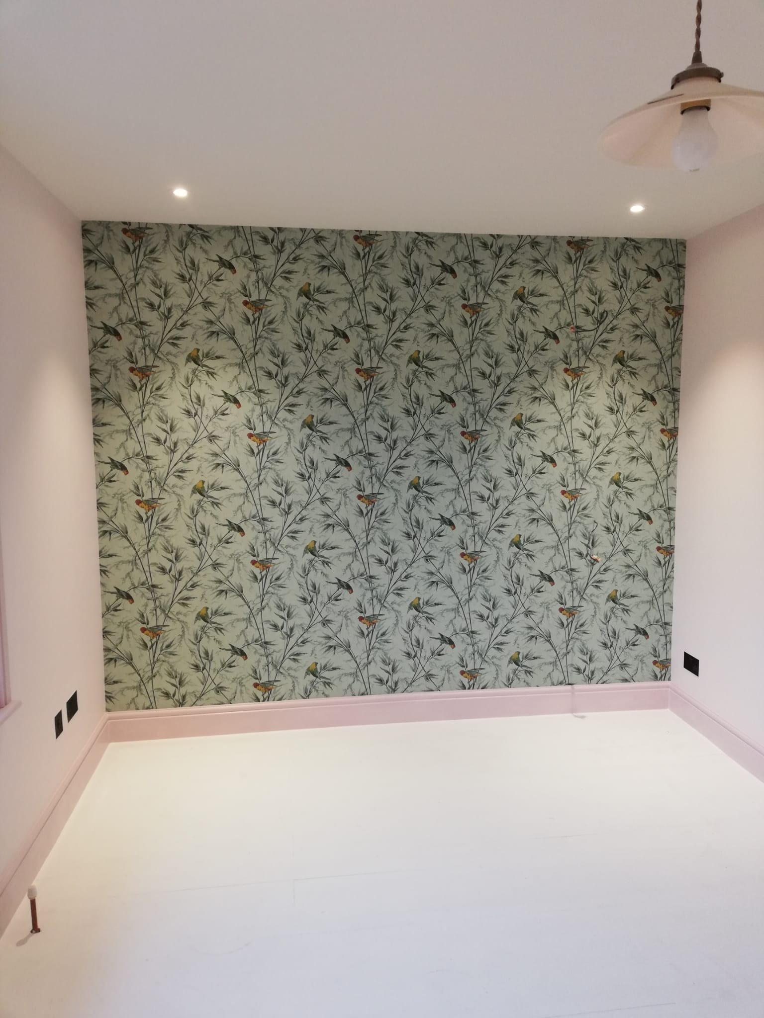 Pusty pokój po remoncie z nową tapetą w motywy roślinne i ptasie, pomalowanymi na różowo ścianami i listwami przypodłogowymi, oraz odnowioną białą podłogą.