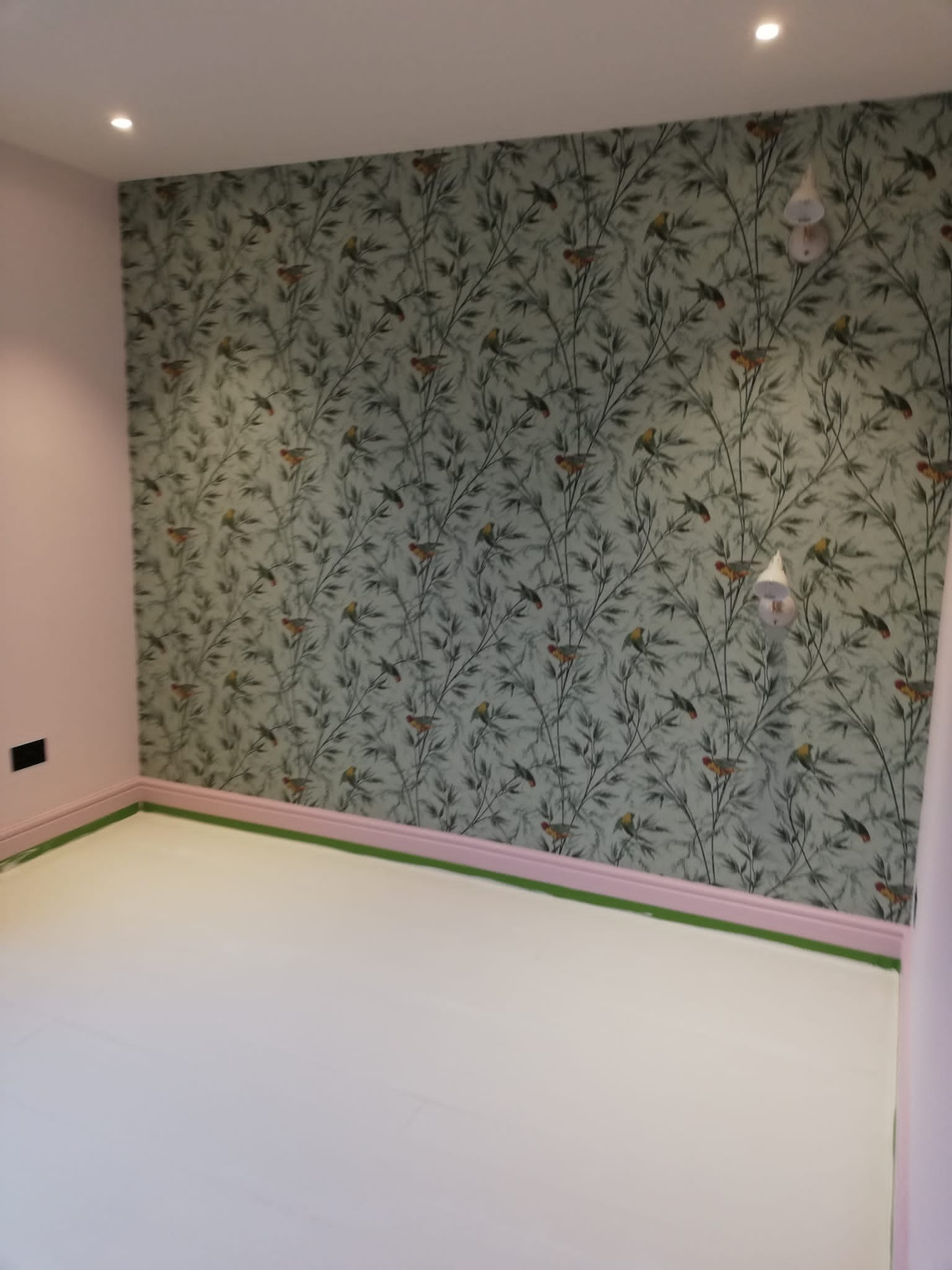 Pomalowane na różowo ściany i listwy przypodłogowe w pokoju z tapetą w motywy roślinne i ptasie, świeżo położona podłoga zabezpieczona taśmą malarską.