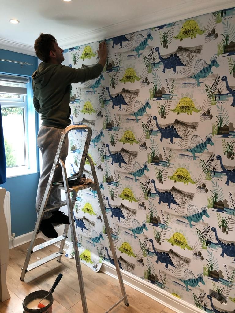 Mężczyzna na drabinie w Bystrej nakleja tapetę w dinozaury na ścianie w pokoju z niebieskimi ścianami i oknem.
