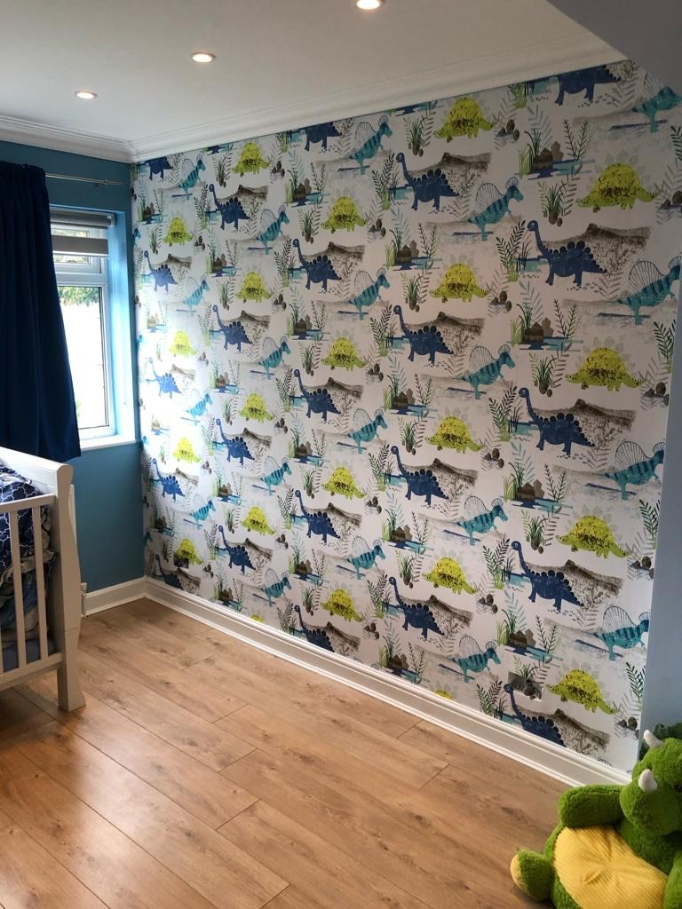 Pokój dziecięcy z drewnianą podłogą, ścianą pokrytą tapetą w dinozaury i fragmentem łóżeczka z niebieską pościelą w tle, oświetlony punktowymi lampami sufitowymi.