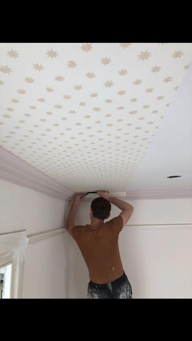 Mężczyzna w brązowej koszulce precyzyjnie maluje pędzlem krawędź sufitu z motywem gwiazd w pokoju z listwami przysufitowymi w kolorze pudrowego różu.