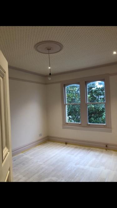 Jasne, odświeżone wnętrze pokoju z parkietem, pomalowanymi ścianami i ozdobnym sufitem z rozetą, widok na zieleń za oknem.