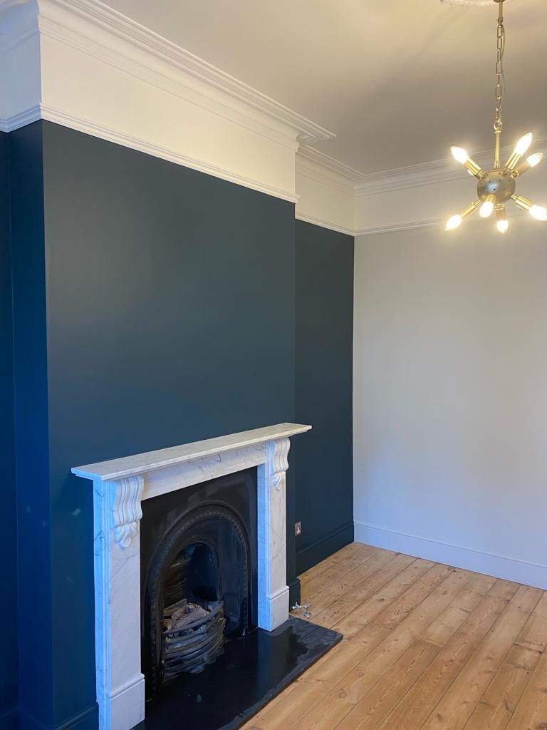 Pokój po remoncie: ściany pomalowane na granatowo i biało, marmurowy kominek z żeliwnym wkładem, drewniana podłoga, nowoczesna lampa sufitowa.