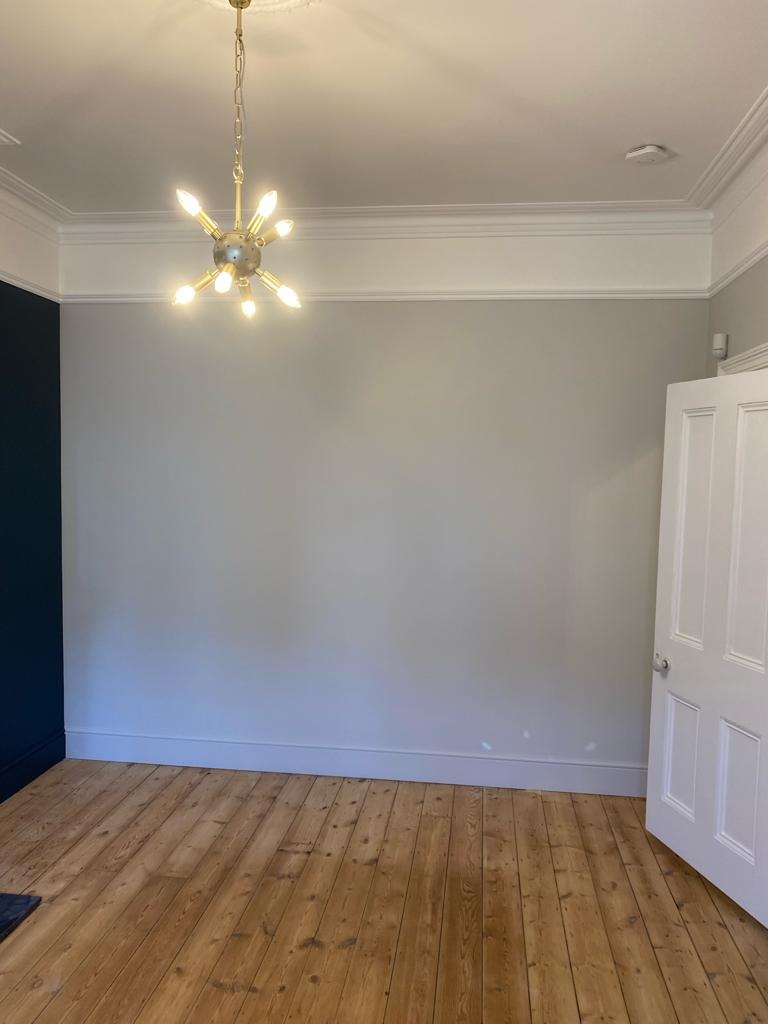 Pomalowane na szaro wnętrze pokoju z drewnianą podłogą, widoczny fragment ściany pomalowany na granatowo, biała listwa przypodłogowa i sufitowa, oraz designerska lampa sufitowa w stylu retro.