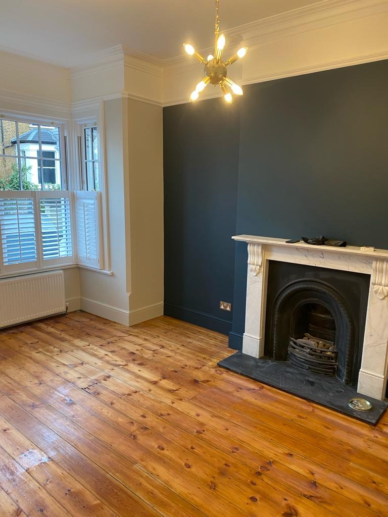 Wnętrze pokoju po odświeżeniu: ciemnogranatowa ściana z marmurowym kominkiem, odnowiona drewniana podłoga i nowoczesna lampa w stylu atomowym.