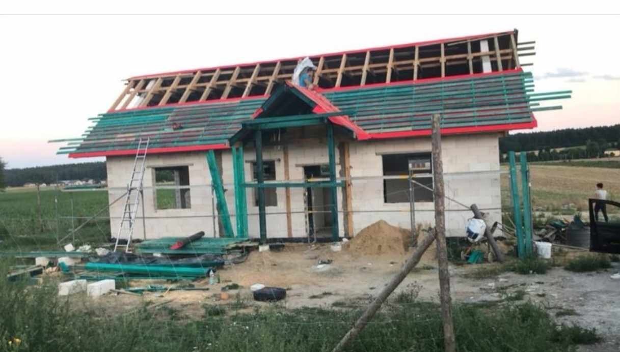 Budowa domu jednorodzinnego w trakcie montażu więźby dachowej i deskowania, widoczne pustaki, niezadaszony ganek, drabina oparta o ścianę, teren budowy z lekkim nieporządkiem.