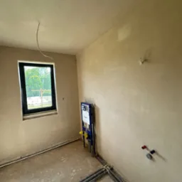 Wnętrze po tynkowaniu maszynowym: gładkie ściany w odcieniach beżu, okno z widokiem na zieleń, instalacja sanitarna i rury przy podłodze. Widać świeżo położony tynk.