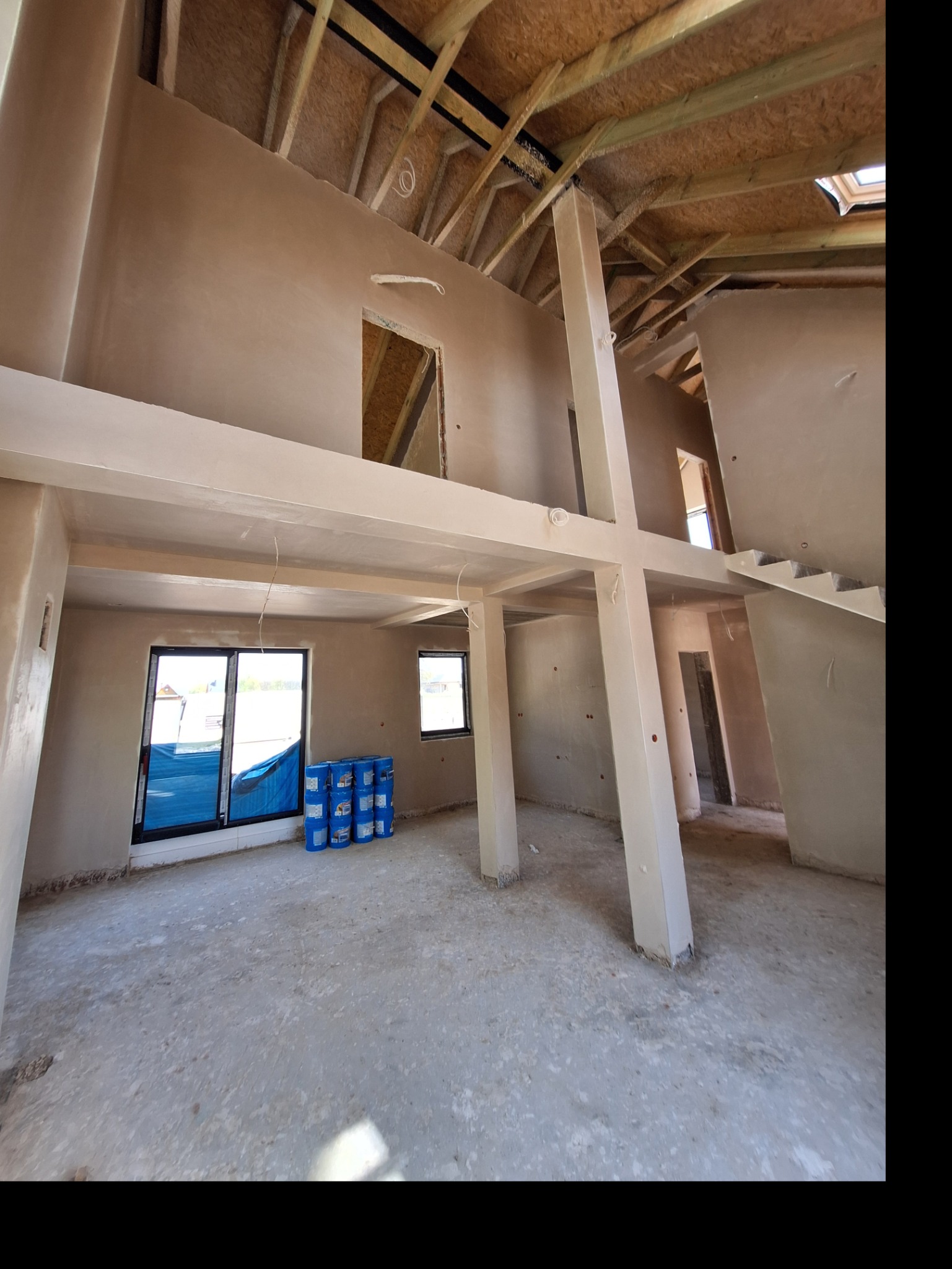 Surowe wnętrze domu po tynkowaniu maszynowym, widoczne ściany, słupy i drewniana konstrukcja dachu. Na podłodze stoją niebieskie wiadra z materiałami.