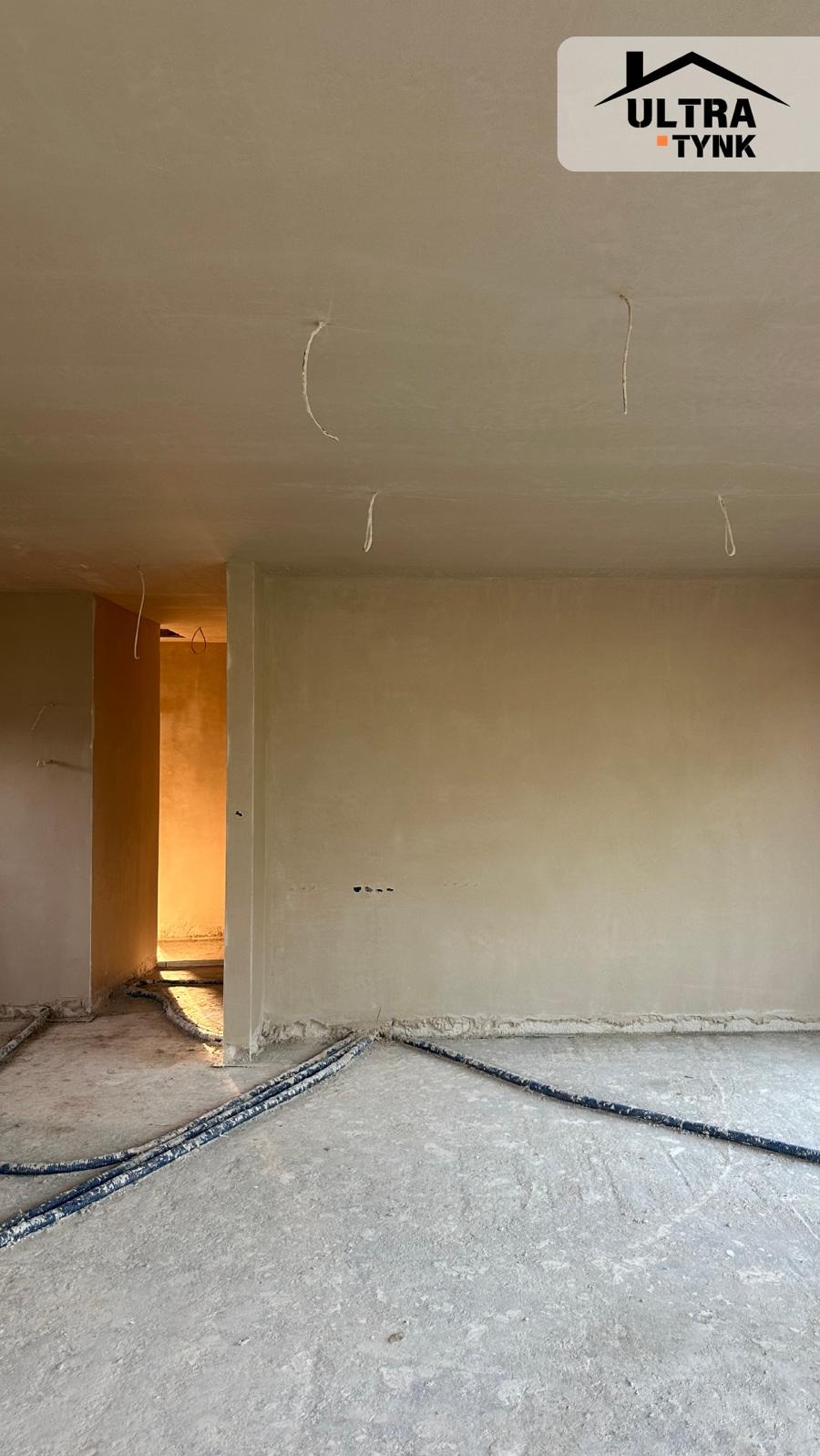 Wnętrze po tynkowaniu maszynowym: gładkie ściany, wystające przewody elektryczne z sufitu, rury na podłodze. Widoczne detale wykończenia i przygotowanie do dalszych prac.