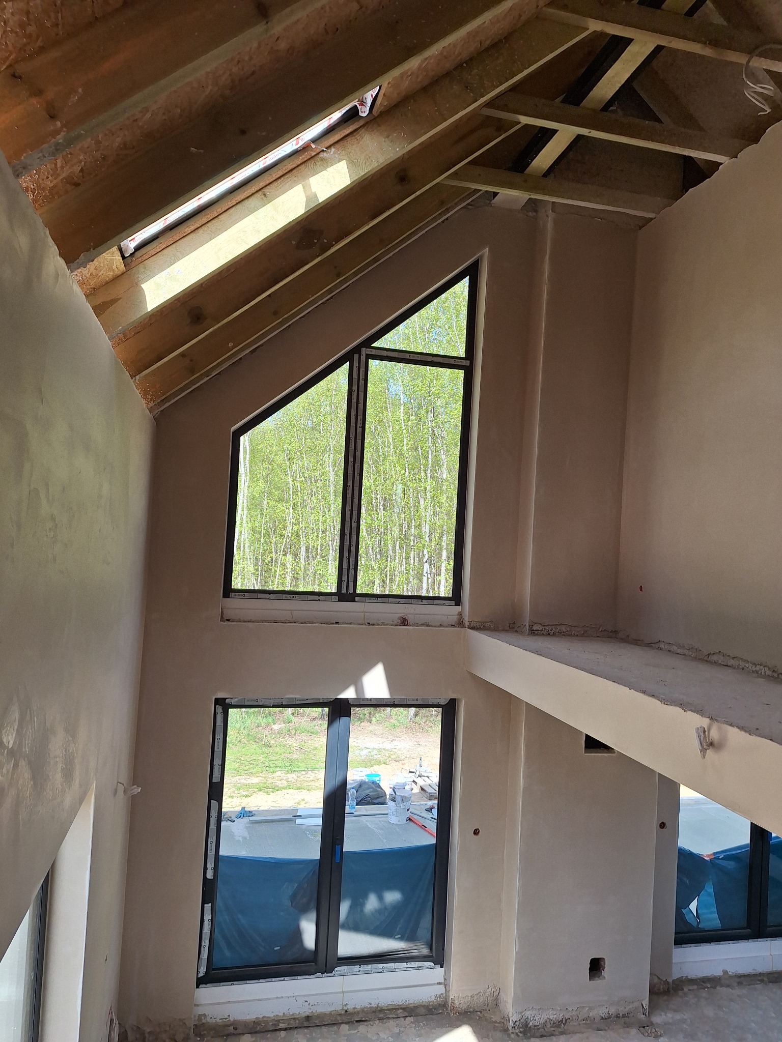 Wnętrze domu w trakcie prac tynkarskich: surowe ściany z oknami o nietypowych kształtach, drewniana konstrukcja dachu, widok na zieleń za oknem.
