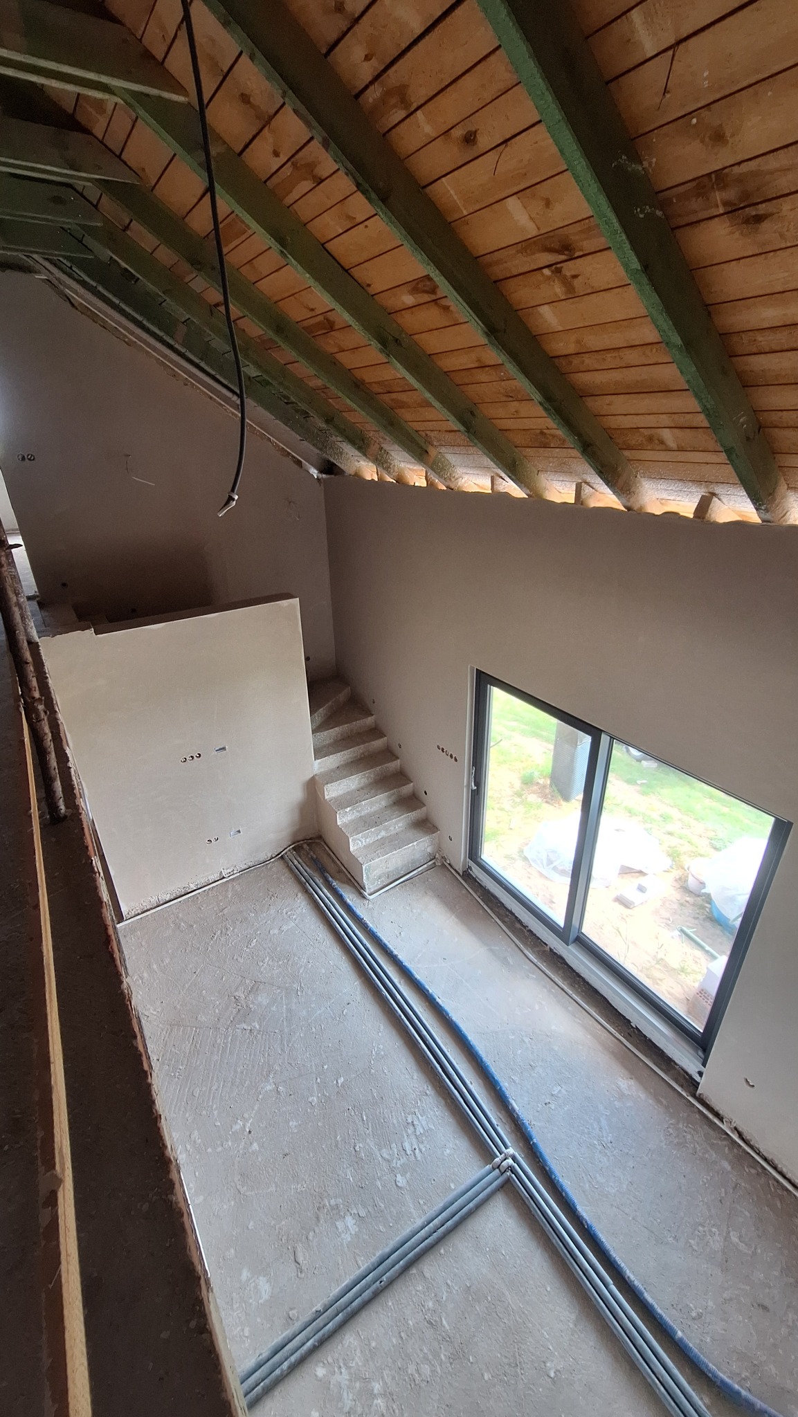 Wnętrze w trakcie prac wykończeniowych: ściany pokryte tynkiem, widoczne przewody na podłodze, drewniany strop. Schody w surowym stanie, duże okno z widokiem na zewnątrz.