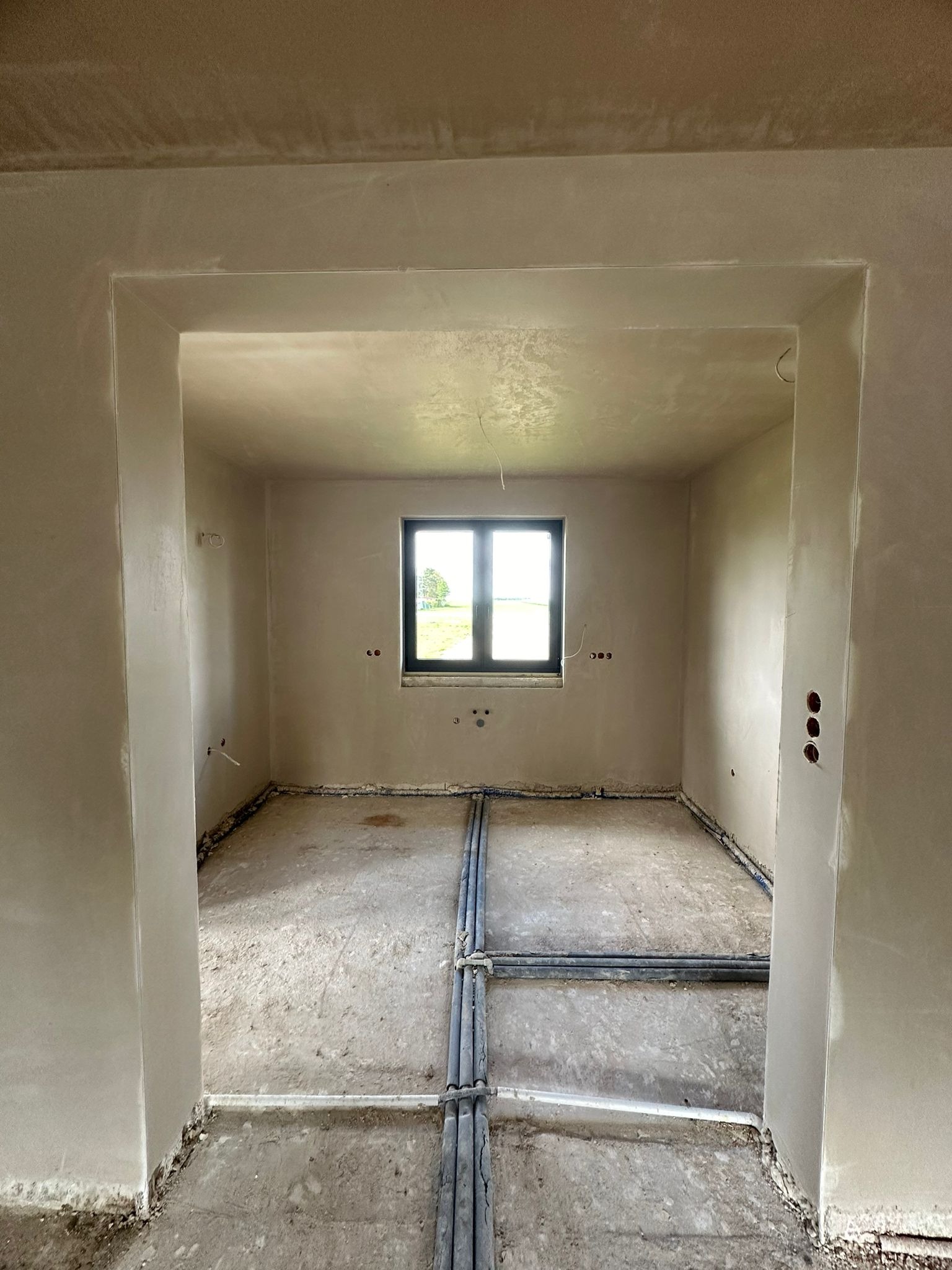Wnętrze po aplikacji tynków maszynowych: gładkie ściany, widok przez otwór drzwiowy na okno z zielenią na zewnątrz. Rury instalacji na podłodze.