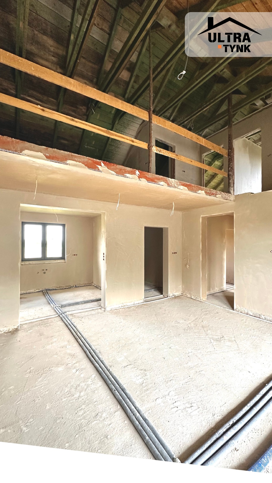 Wnętrze domu po nałożeniu tynków z widocznymi rurami instalacji grzewczej w podłodze, surowym drewnianym stropem i balustradą. Widoczne ślady po tynkowaniu.