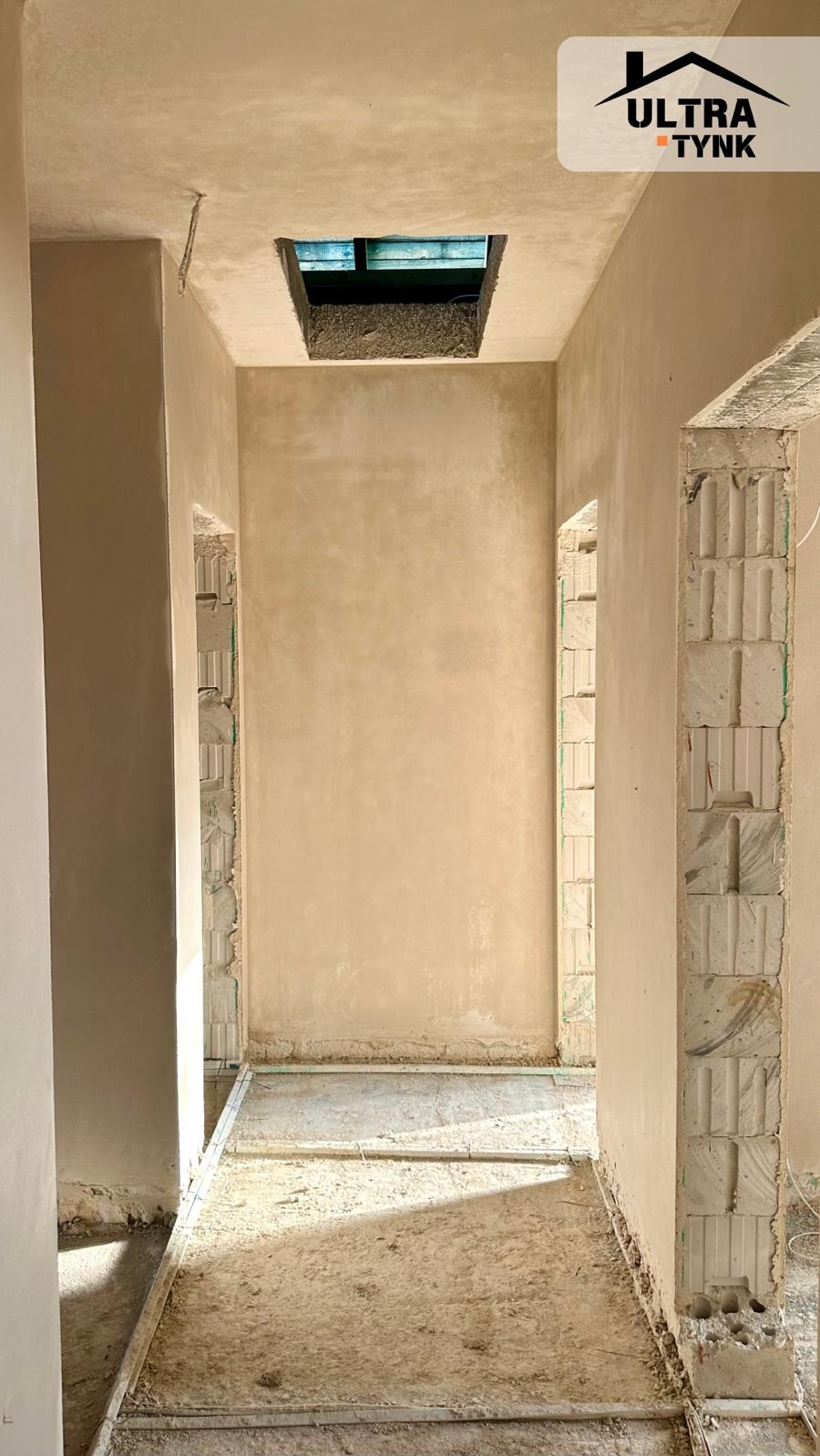Korytarz po tynkowaniu maszynowym, widoczne ściany i sufit pokryte świeżym tynkiem, w suficie otwór na okno dachowe, na podłodze resztki materiałów.