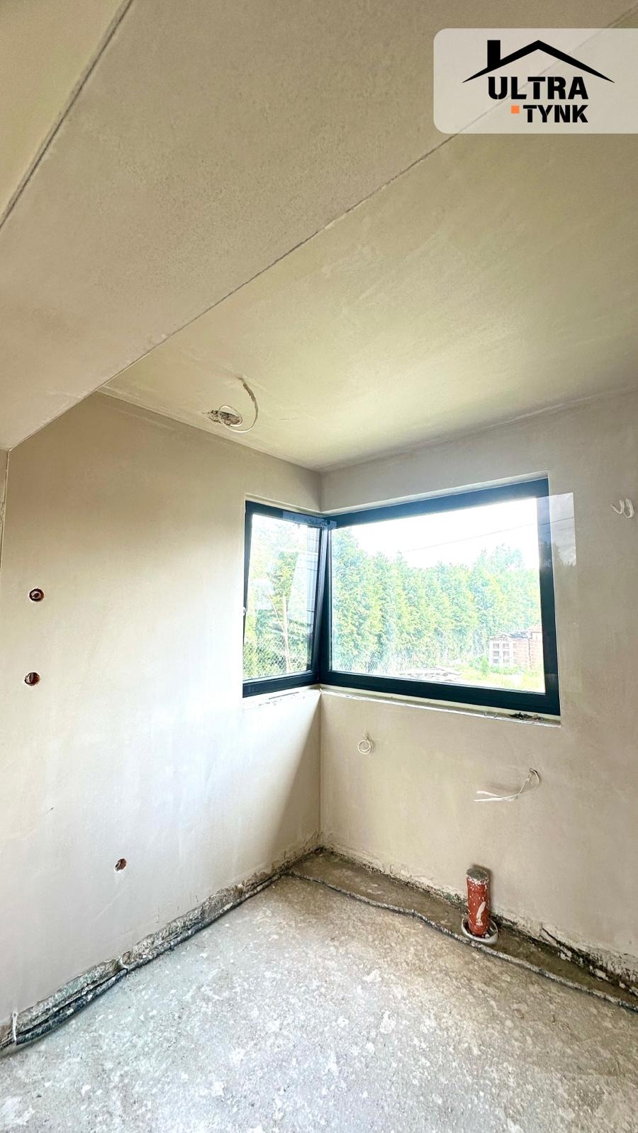 Wnętrze pokoju po nałożeniu gładzi, z widocznym oknem narożnym i wystającymi elementami instalacji elektrycznej. Ściany w jasnym odcieniu szarości.