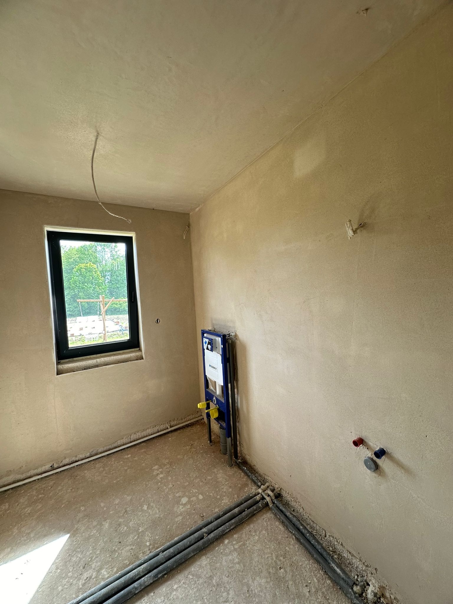 Wnętrze po tynkowaniu maszynowym: gładkie ściany w odcieniach beżu, okno z widokiem na zieleń, instalacja sanitarna i rury przy podłodze. Widać świeżo położony tynk.