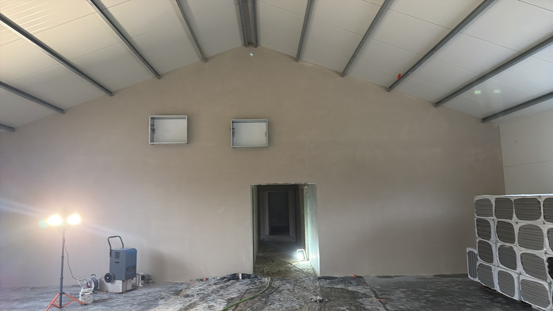 Wnętrze po nałożeniu gładzi: ściany w odcieniach beżu, widoczne otwory wentylacyjne, sprzęt budowlany i materiały. Oświetlenie halogenowe podkreśla strukturę powierzchni.