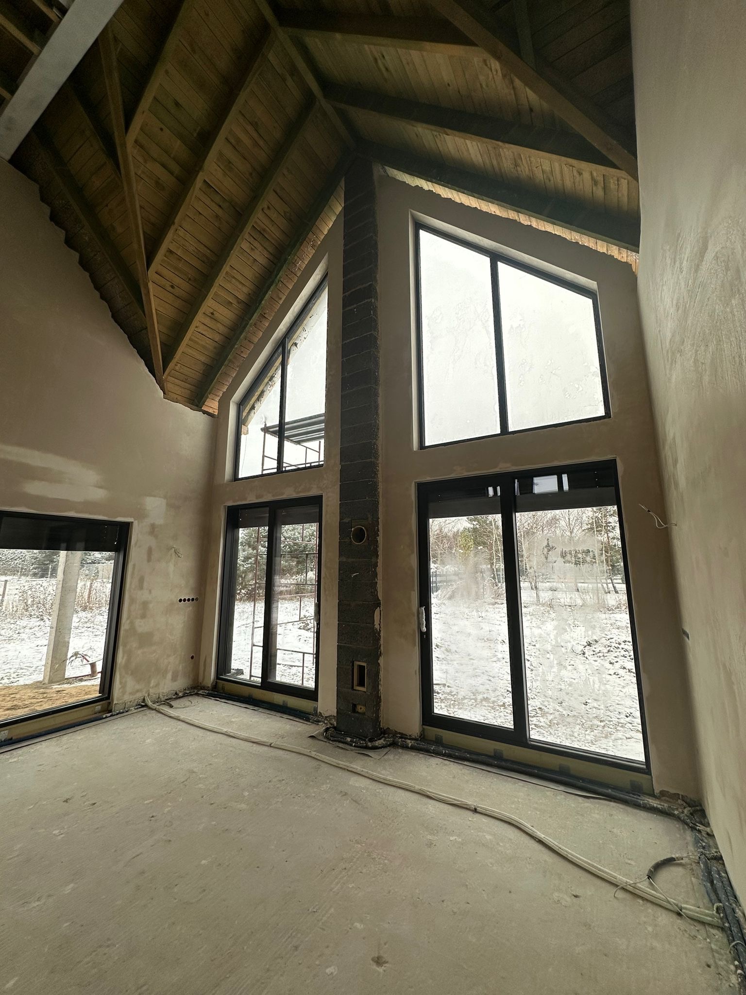 Wnętrze domu w trakcie prac wykończeniowych: surowe tynki na ścianach, drewniany strop, duże okna z widokiem na zimowy krajobraz. Widoczne przewody instalacji.