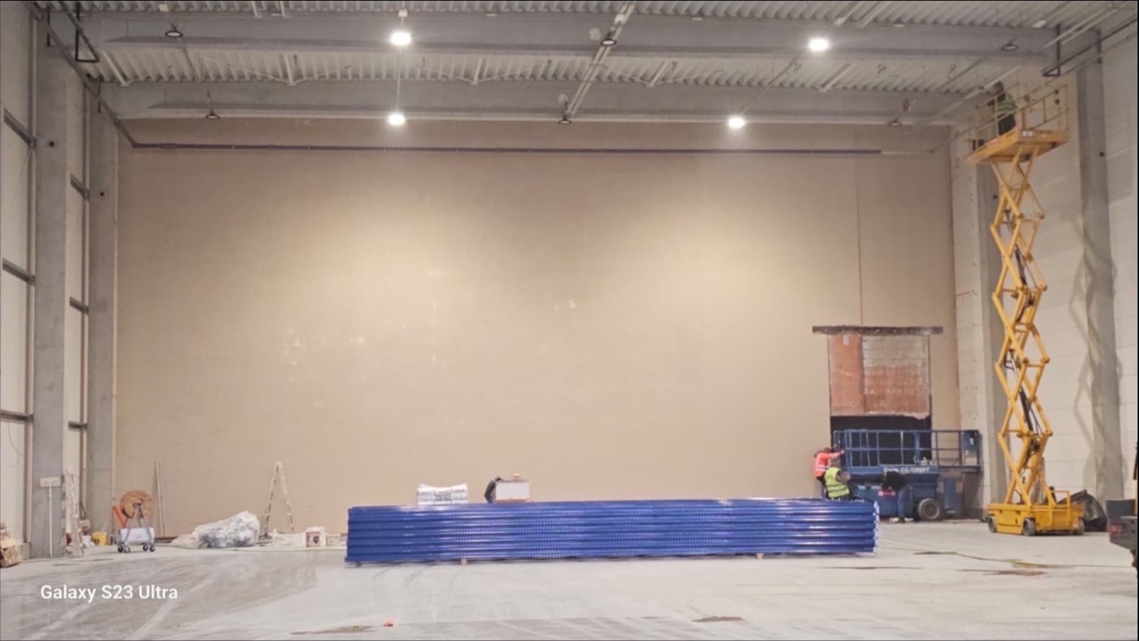 Wnętrze hali w trakcie prac wykończeniowych. Ściana po nałożeniu tynku, podnośnik z pracownikiem przy otworze okiennym, materiały budowlane na podłodze.