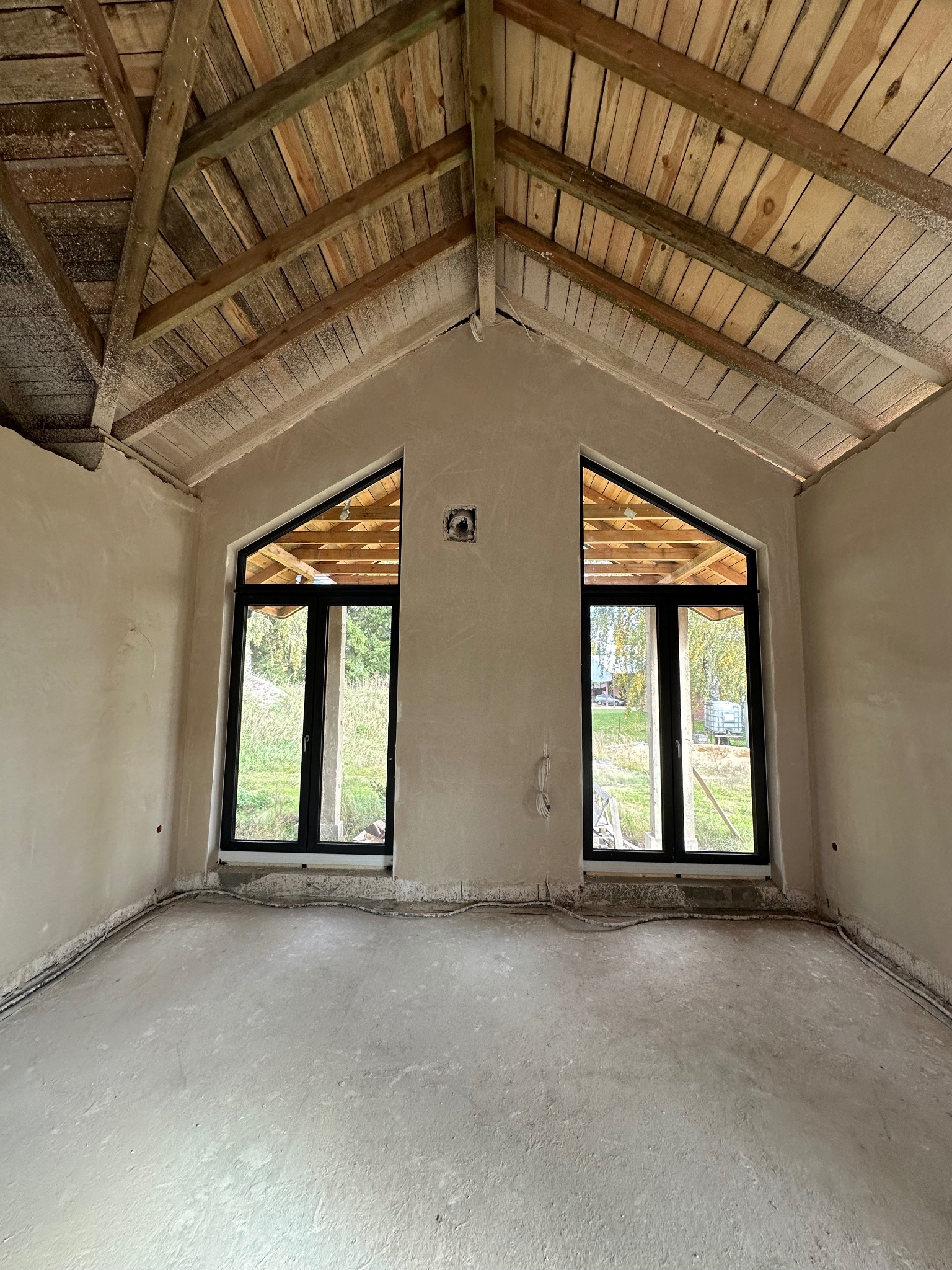 Wnętrze domu po nałożeniu tynków, widoczne dwa okna o trójkątnym kształcie u góry, drewniany strop i szare ściany. Na podłodze widać wystające kable.