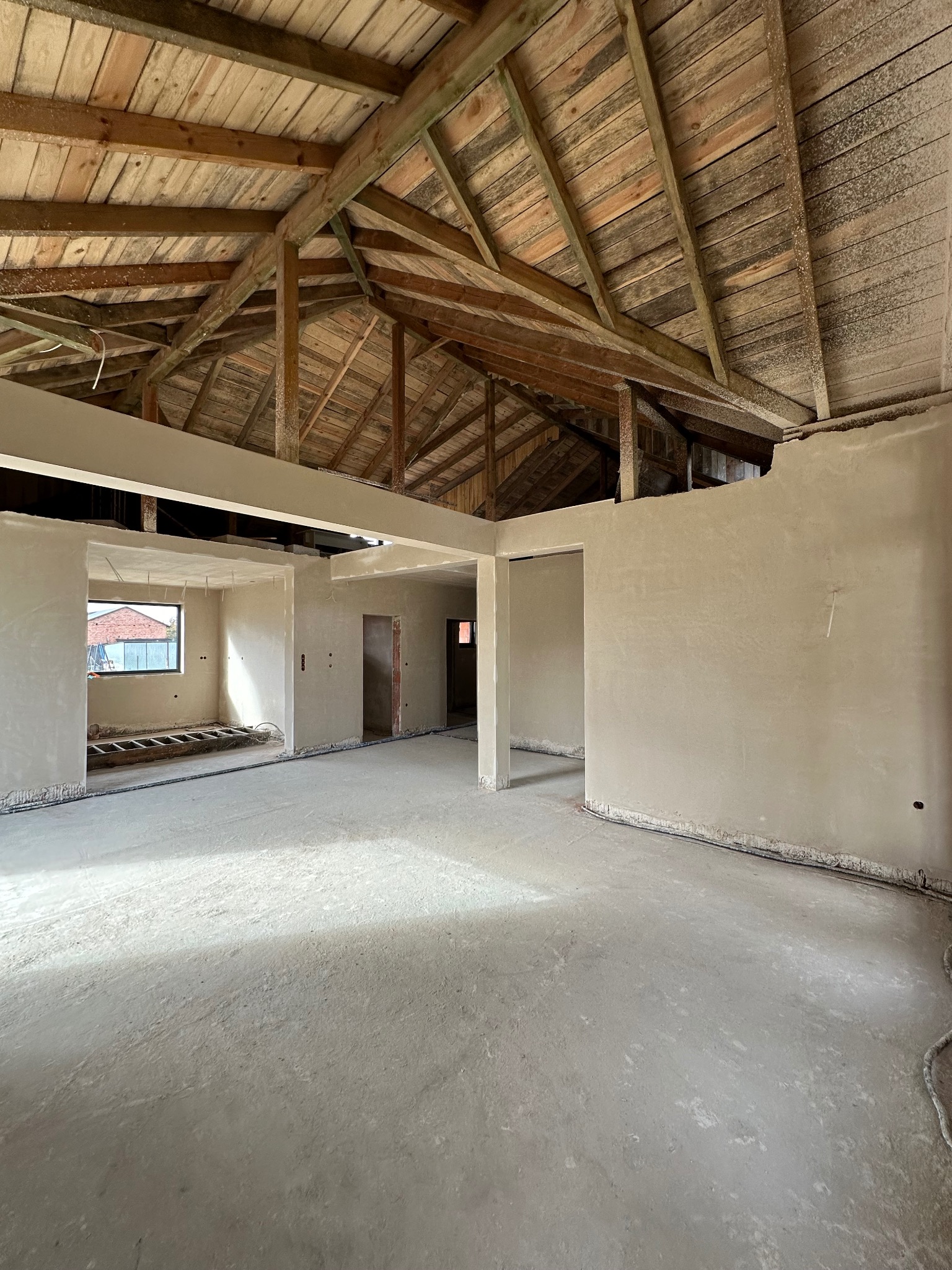 Wnętrze domu w trakcie prac wykończeniowych: surowe ściany po nałożeniu tynku maszynowego, drewniana konstrukcja dachu, widoczne przewody elektryczne przy podłodze.