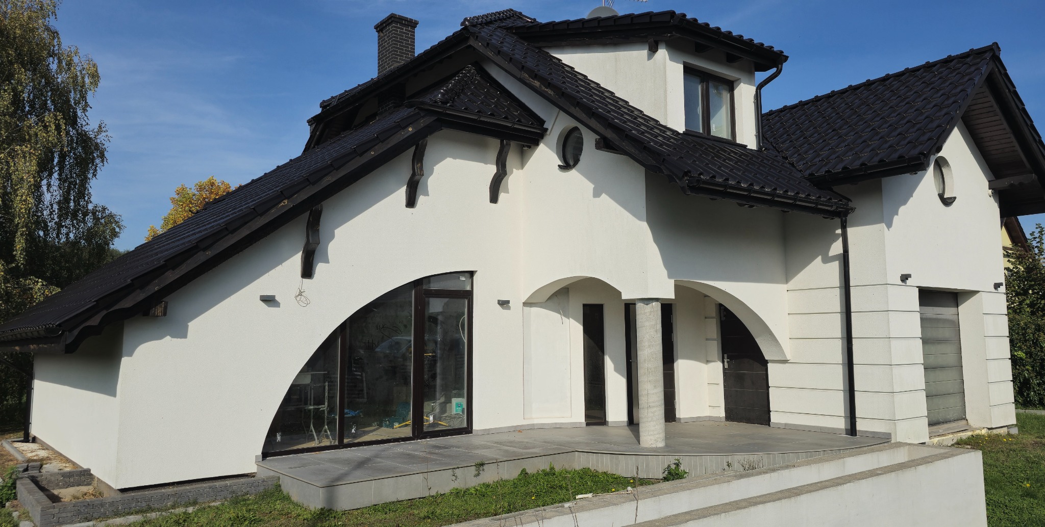 Nowoczesny dom murowany z białym tynkiem, czarną dachówką i charakterystycznym łukowym oknem na parterze, widok z zewnątrz w słoneczny dzień.