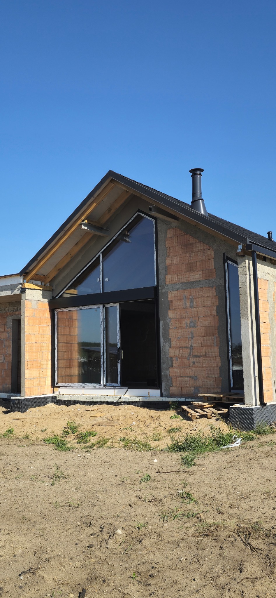 Surowy stan domu z czerwonej cegły z dużym oknem szczytowym i kominem, widoczne elementy konstrukcyjne i podłoże z piasku.