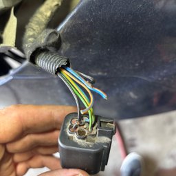 Hubert Bielecki - Uszkodzona wiązka elektryczna samochodu z widocznymi przewodami w kolorach: niebieskim, zielonym i pomarańczowym, trzymana w dłoni, z fragmentem nadkola w tle.